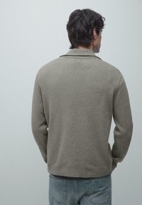 Mann mit dunklen Haaren, der einen grauen Pullover mit langen Ärmeln und eine blaue Jeans trägt, steht mit den Händen in den Taschen, von hinten betrachtet.