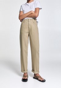 Pantalones de talle alto color beige con dobladillos enrollados, hechos de una tela de algodón grueso, combinados con una camiseta blanca básica y chanclas negras.