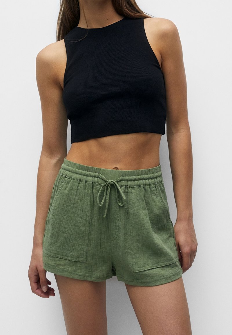 Femme portant un top court noir sans manches et un short vert à cordon, debout devant un fond uni.