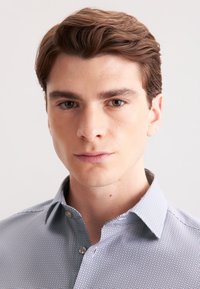 Camicia azzurra chiara con un motivo a micro-check. Presenta un colletto alla francese e una chiusura con bottoni sul davanti. Il tessuto appare liscio e leggero.