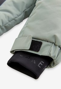 Mănușă izolată verde cu o manșetă ajustabilă cu velcro, având un căptușeală interioară elastică neagră, embosată cu text. Textură moale și netedă.