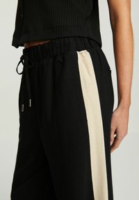 Zwarte broek met een contrasterende beige zijkantstreep, elastische tailleband en verstelbaar trekkoord. Gemaakt van zachte stof met een ontspannen pasvorm.