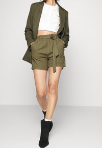 Blazer verde oliva e pantaloni corti coordinati con cintura. Camicia bianca corta con colletto arricciato. Stivaletti neri alla caviglia. Tessuto dalla consistenza liscia.