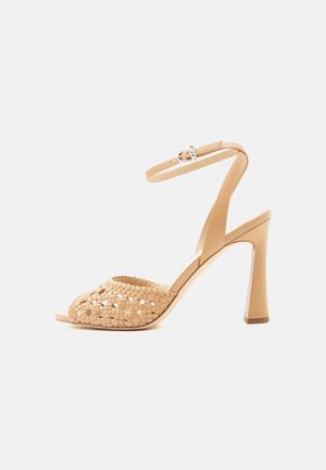Sandalo beige con tacco alto, cinturino intrecciato sulla parte anteriore e cinturino alla caviglia con fibbia su sfondo chiaro uniforme.