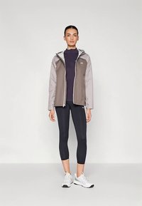 Lekka szara kurtka z kapturem, zapinana na zamek, z bocznymi kieszeniami, nałożona na fioletową koszulę i czarne legginsy, w zestawie z białymi trampkami.