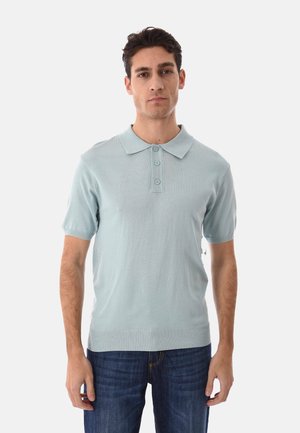 Giovane uomo che indossa una polo a maniche corte azzurro chiaro e jeans blu scuro, in piedi davanti a uno sfondo bianco neutro.