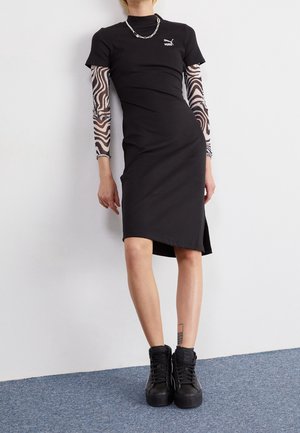 Mujer que lleva un vestido negro Puma con mangas largas de estampado de cebra debajo, zapatillas negras de plataforma y un collar de cadena plateada.