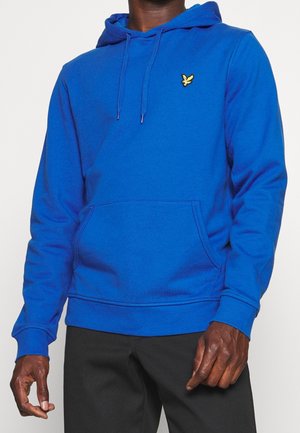 Man wearing a bright blue hoodie with front pocket and yellow bird logo on chest, paired with black pants against a plain background.