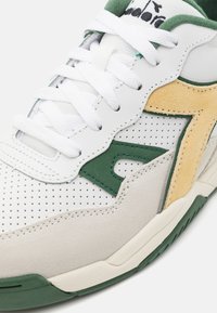 Sneaker in pelle bianca con accenti in suede verde e giallo, tomaia perforata, lacci piatti bianchi e suola in gomma testurizzata.