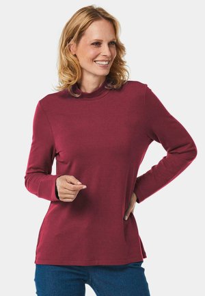 Bordeaux langemouwen top met een hoge hals en zijsplitten. Zachte stof met een gladde textuur, gecombineerd met blauwe jeans.