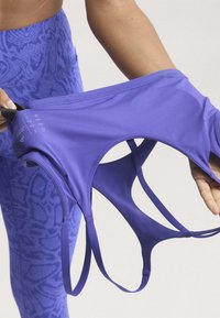 Personne étirant une brassière de sport violette à multiples fines bretelles, portant un legging bleu à motifs, sur un fond clair.