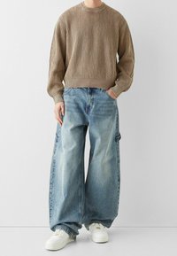 Hnědý žebrovaný pletený svetr, volný střih. Světle modré oversized džíny s efektem vyprání, detail na bočních kapsách. Bílé tenisky doplňují outfit.