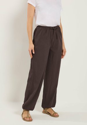 Mujer vistiendo pantalones sueltos marrón oscuro con cordón, camiseta blanca de manga corta y sandalias marrones, de pie contra un fondo liso.