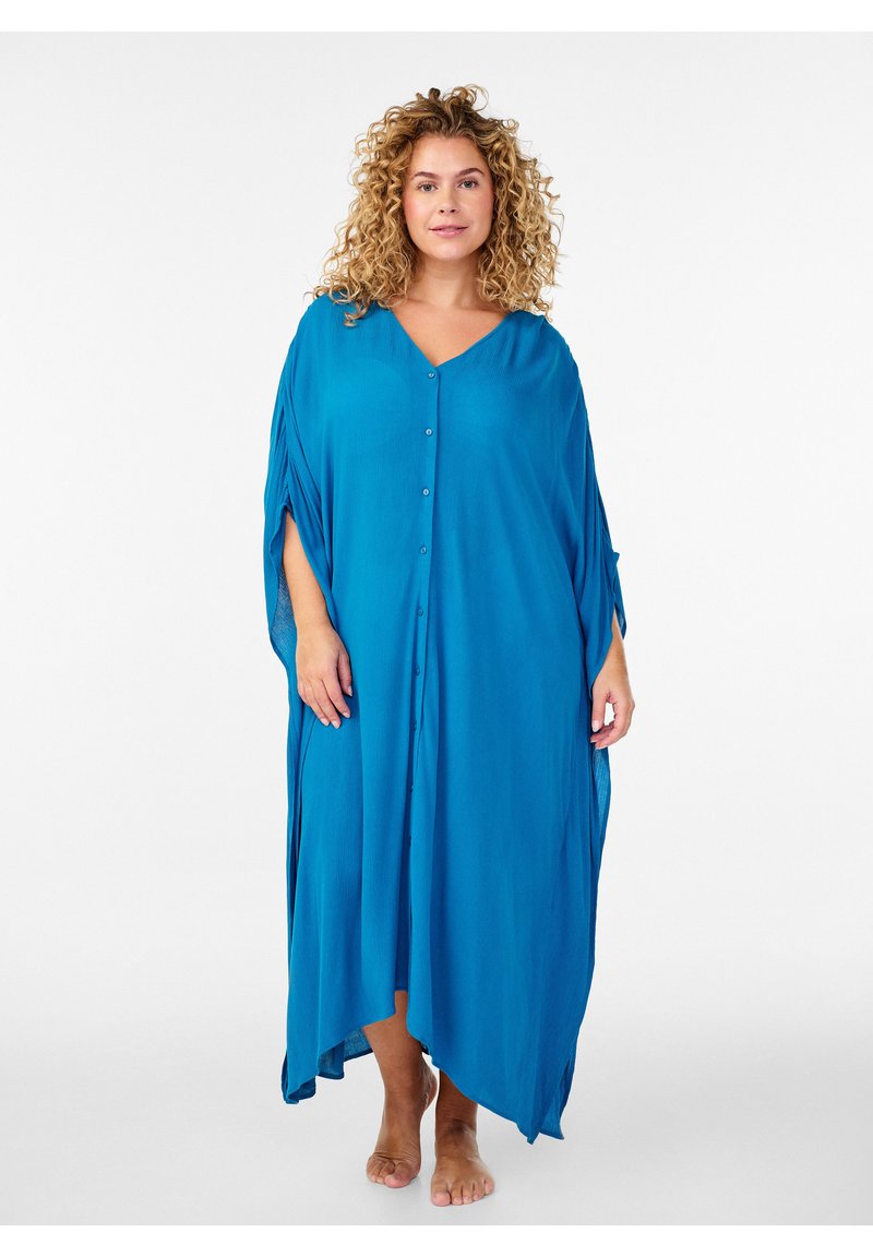 Kobieta o kręconych blond włosach, ubrana w luźną, jasnoniebieską, zapinaną na guziki sukienkę-kaftan, stojąca boso na białym tle.