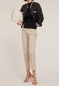 Czarna kurtka puchowa bomberka z beżowymi akcentami, prążkowany czarny golf, beżowe tapered trousers oraz beżowe sandały na obcasie, trzymając kremową torebkę.