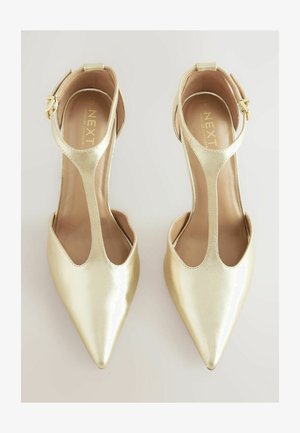 Zelta metāliskas augstpapēžu kurpes ar norādītiem pirkstiem un T-siksnas dizainu. Iekšējā oderes virsma ir gluda, ar minimālu šuvju detaļu.