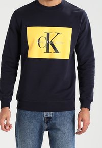 Sudadera azul marino con un logotipo rectangular amarillo que presenta "Calvin Klein Jeans" y "CK" en negro, fabricada en algodón y con diseño de cuello redondo.
