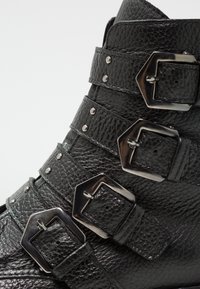 Černé kožené kotníkové boty s několika vodorovnými pásky se stříbrnými čtvercovými sponami a hřebíky, s texturovaným povrchem.