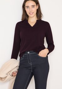 Langemouw, diep bordeaux top met een V-hals ontwerp, gecombineerd met donkerblauwe jeans met contrasterende stiksels, gedragen met een gezellige beige jas.