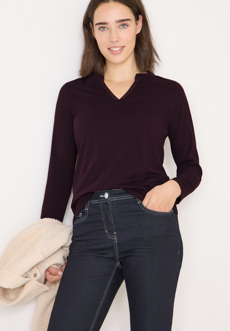 Langemouw, diep bordeaux top met een V-hals ontwerp, gecombineerd met donkerblauwe jeans met contrasterende stiksels, gedragen met een gezellige beige jas.