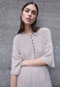 Γυναίκα με βρεγμένα σκούρα μαλλιά που φοράει μπεζ πλεκτό φόρεμα με μανίκια μέχρι τον αγκώνα και πέρλες κουμπιά, στέκεται μπροστά σε γκρι τοιχο με υφή.