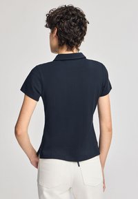 Marinblå kortärmad polo med krage, med rak fåll och en liten sidoven, bärs med ljusa byxor.