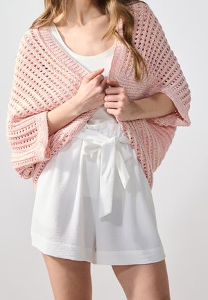 Žena má na sobě světle růžový pletený šál přes bílý top bez rukávů a bílé šortky s vysokým pasem a zavazovacím pásem.