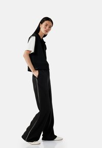 Un gilet en tricot noir porté sur un t-shirt blanc, associé à un pantalon large noir avec des bordures blanches, et des chaussures de couleur crème.