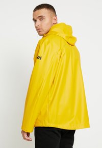 Giacca impermeabile gialla con cappuccio, realizzata in materiale lucido e liscia. Presenta una fessura laterale e un logo nero sulla manica sinistra.