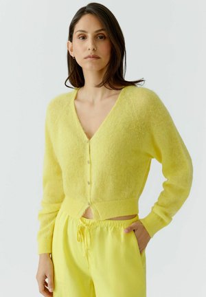 Femme portant un cardigan jaune duveteux boutonné et un pantalon jaune à cordon, debout avec une main dans la poche, devant un fond uni.