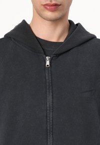 Black zip-up hoodie featuring a front zipper, a drawstring hood, and a subtle logo print on the left chest area, with a soft texture.