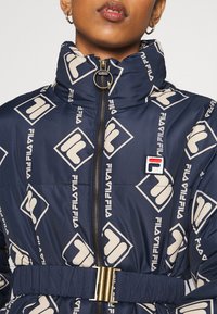 Granatowa kurtka puchowa z powtarzającym się beżowym wzorem logo FILA, z wysokim kołnierzem, zamek błyskawiczny z przodu oraz paskiem w talii z złotymi elementami.