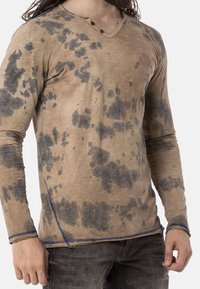 Camicia beige a maniche lunghe con motivo a macchie grigio scuro; presenta un collo a V, due bottoni decorativi e cuciture blu a contrasto sull'orlo.
