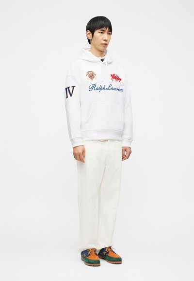 Νεαρός άνδρας φορώντας λευκό φούτερ Ralph Lauren με κουκούλα, λευκό παντελόνι και πολύχρωμα καφέ, πράσινα και μπλε δερμάτινα παπούτσια με κορδόνια, στέκεται σε λευκό φόντο.