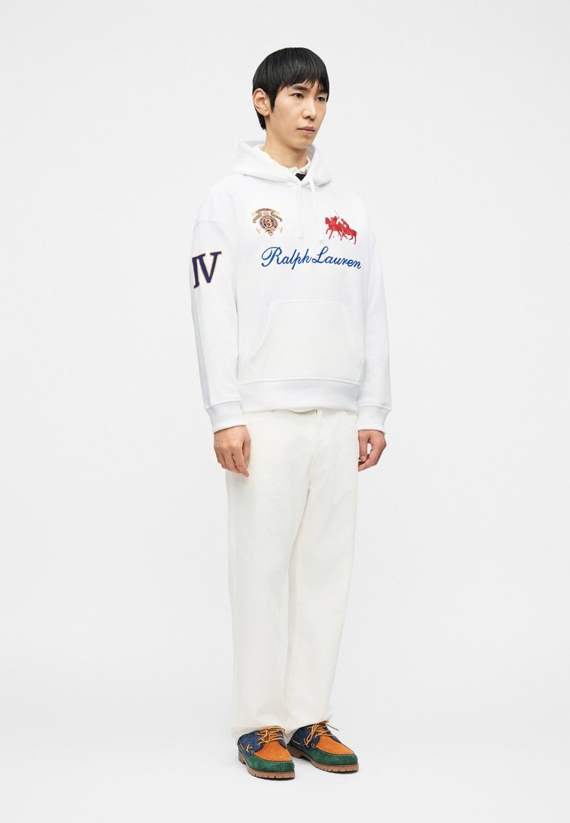 Jeune homme portant un sweat à capuche blanc Ralph Lauren, un pantalon blanc et des chaussures à lacets multicolores marron, vert et bleu, debout sur un fond blanc.