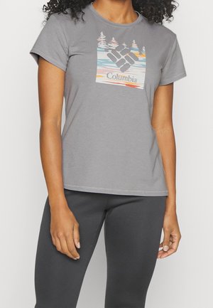 Femme portant un t-shirt Columbia gris à manches courtes avec un motif coloré de montagnes et d’arbres et un pantalon gris foncé.