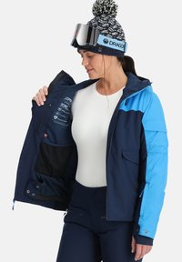 Marineblaue Skijacke mit Kapuze, ausgestattet mit einem Reißverschluss vorne, zwei Außentaschen sowie einer Innenkennzeichnung mit Pflegehinweisen und Merkmalen.