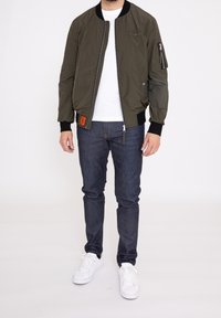 Olivgrön bomberjacka med ribbade mudd, dragkedjefickor och en orange etikett, tillsammans med mörka denimjeans och vita sneakers.