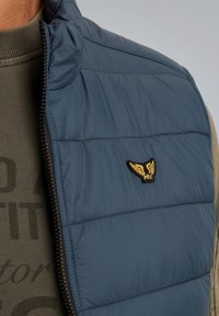 Marinblå vadderad väst med hög krage, med horisontell sömnad och en guld broderad logotyp. Lagerad över en grön texturiserad skjorta.