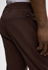 Bruine broek van gladde stof, met een achterzak met een logo-label en een slanke pasvorm. De textuur lijkt zacht met een nette afwerking.