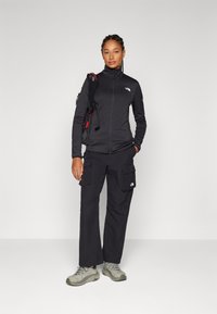 Black zip-up jacket with high collar, paired with black cargo pants featuring side pockets. Grey hiking shoes with textured soles.