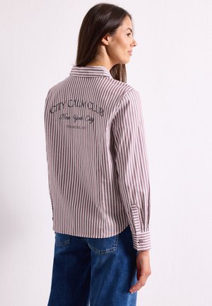 Donna di profilo che indossa una camicia a righe verticali marroni e bianche con testo sulla schiena e jeans blu contro un muro semplice.