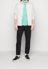 Světle béžová košile na knoflíky přes tyrkysovou košili, doplněná černými kalhotami a černými teniskami s bílými podrážkami. Textura a barvy kontrastují.