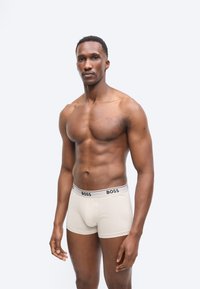 Homme torse nu avec une silhouette athlétique portant un boxer beige avec une ceinture élastique marquée "BOSS", debout devant un fond clair uni.