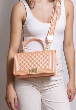 Donna con canottiera bianca e pantaloni tiene una borsa trapuntata color pesca Marc Ellis con tracolla brandizzata attraversando il torso.