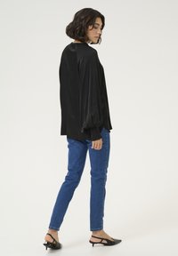 Černá blůza s dlouhým rukávem, balónovými rukávy a pružnými manžetami, kombinovaná s modrými slim-fit džíny. Modelka má na sobě černé podpatkové mule a je otočena mírně stranou.