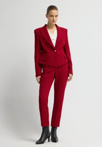 Červený oblek na míru s strukturovaným sakem se zapínáním na jeden knoflík a dvěma předními kapsami, v kombinaci se slim-fit kalhotami a černými kotníkovými botami na podpatku.