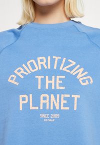 Ljusblå sweatshirt med rund halsringning och raglanärmar. Har stor, texturerad persikofärgad text: "PRIORTERA PLANETEN," och en liten logotyp nedanför.
