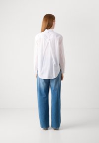 White long-sleeve shirt with curved hem and back pleat detail, paired with light blue wide-leg jeans, featuring a faded texture.