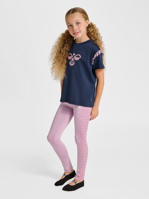 Junges Mädchen mit langen lockigen Haaren, das ein marineblaues Shirt mit Rüschenärmeln und rosa gemusterte Leggings trägt, steht vor einem schlichten Hintergrund.